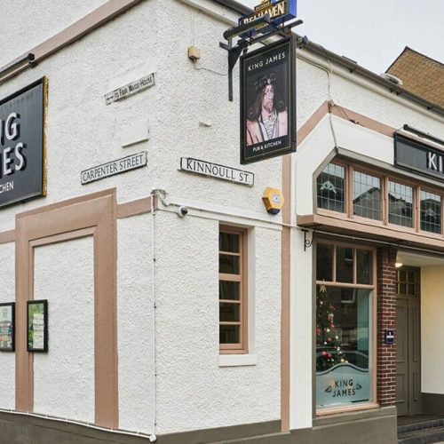 A white pub building in Perth with beige windows and a black sign with a portrait of Scottish King James