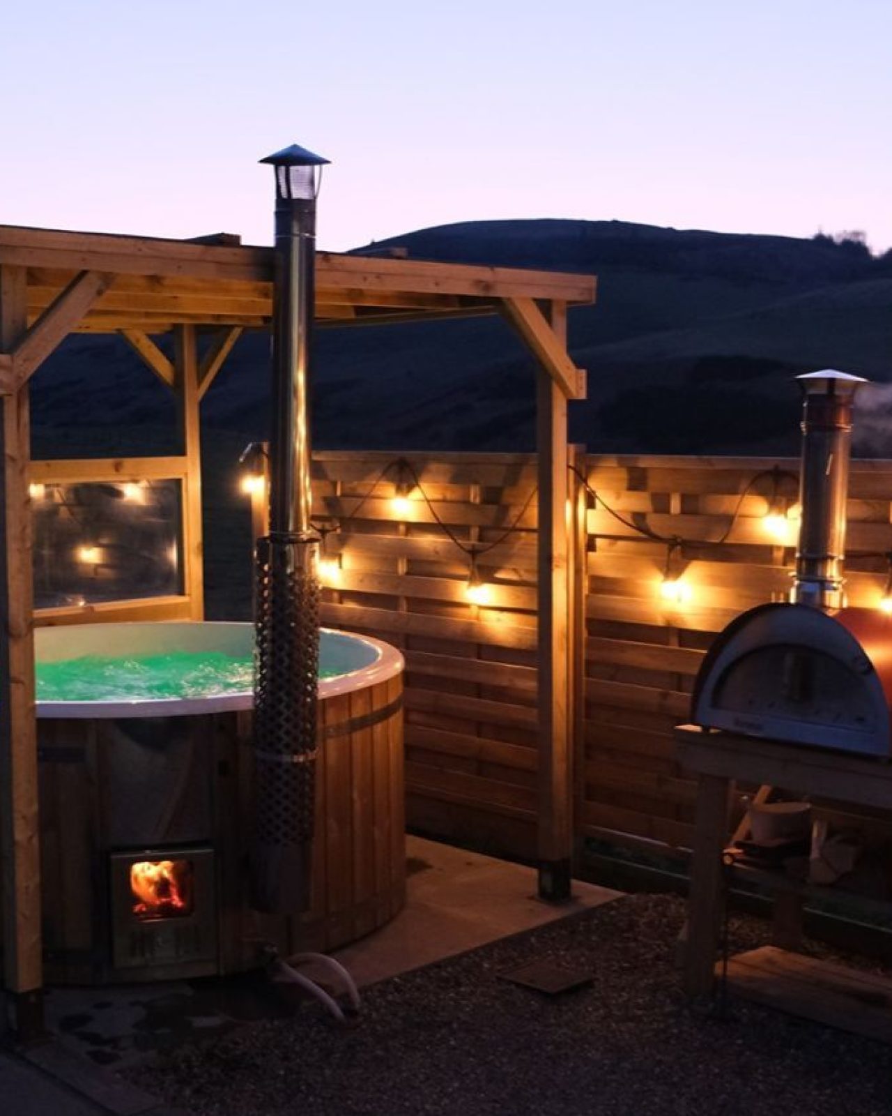 Outdoor hot tub with wooden gazebo and fencing  decorated with fairy lights beside a pizza oven on a wooden table at dusk.
