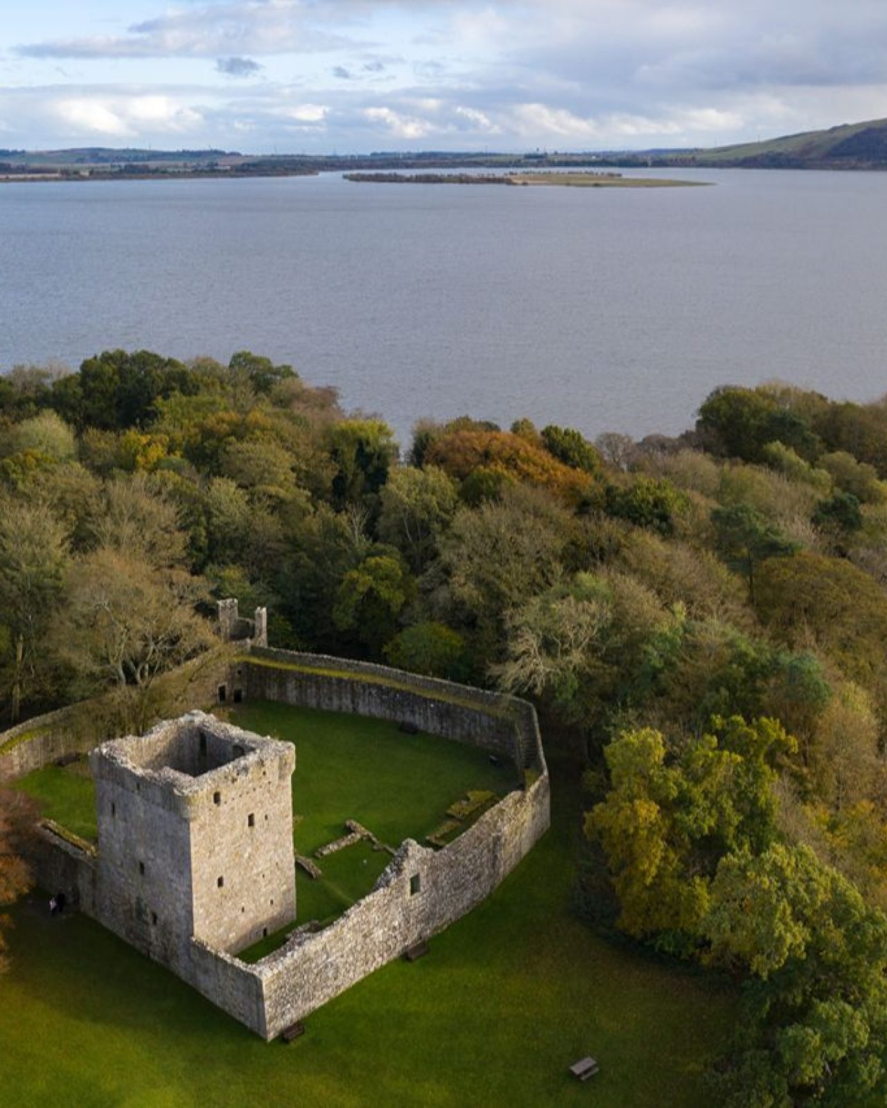 An island with green grass and trees with a old stone turreted building, with a stone wall surrounding, and expanse of water and hills in distance.