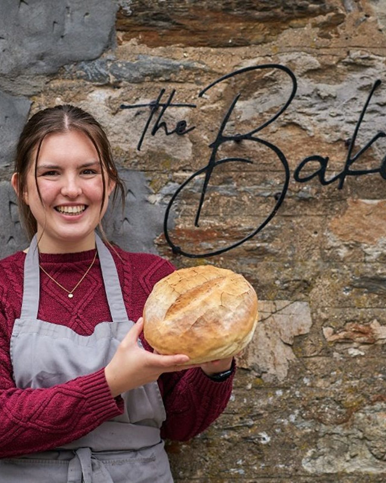 A smiling woman holding a loaf of bread standing next to a black sign which says 'The Bakery' on it.