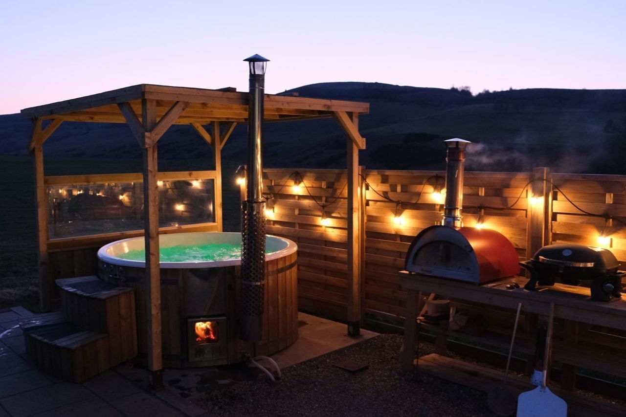 Outdoor hot tub with wooden gazebo and fencing  decorated with fairy lights beside a pizza oven on a wooden table at dusk.