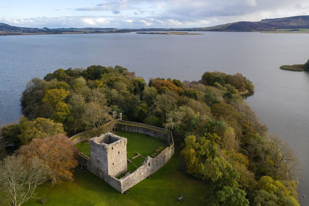 An island with green grass and trees with a old stone turreted building, with a stone wall surrounding, and expanse of water and hills in distance.