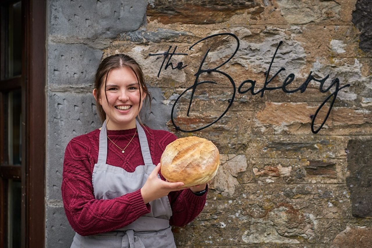 A smiling woman holding a loaf of bread standing next to a black sign which says 'The Bakery' on it.
