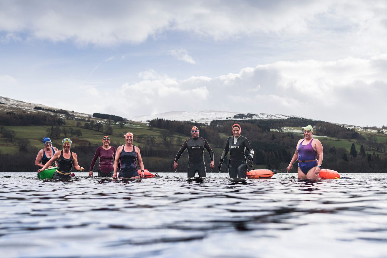 A group of people wearing swimwear standing knee deep in a loch looking very happy.
