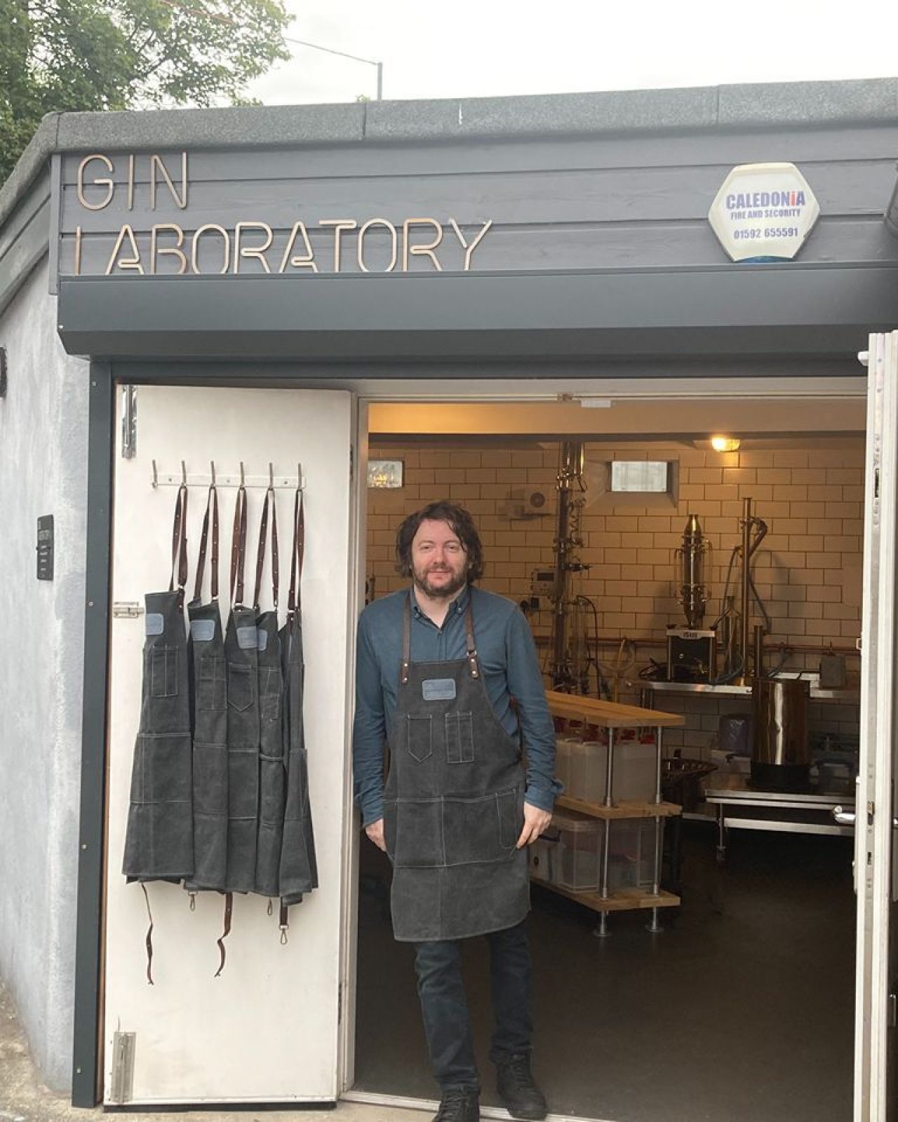 A man wearing a blue shirt and black apron leans on a doorway which opens into a room filled with distillery equipment in Kinross