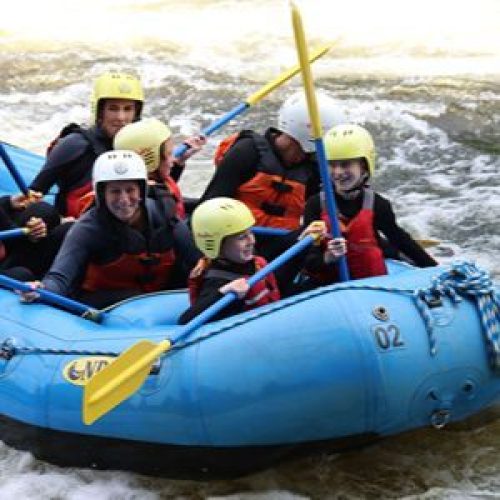 Lots of smiling people holding paddles in a large kayak in fast flowing water.