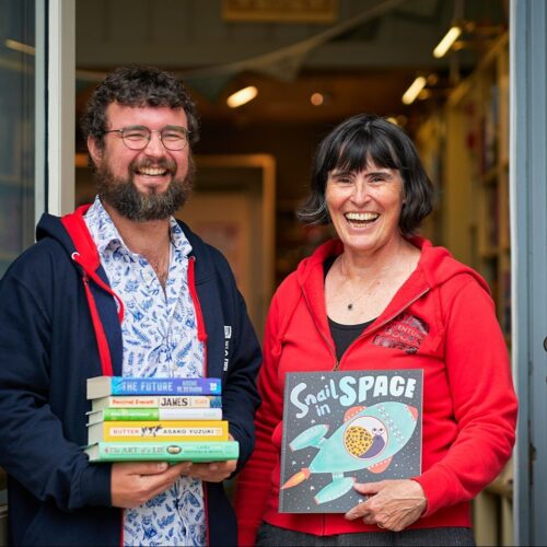 A smiling man wearing a black jacket standing next to a smiling woman wearing a red jacket, both holding books.
