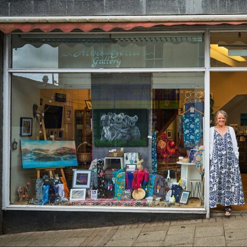 A smiling woman wearing a blue and white dress stands in a doorway next to a very large window displaying artwork and textiles.
