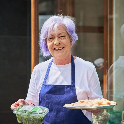 A smiling older woman with purple hair wearing a white shirt and blue apron holds a plate of raw fish and a tub of rocket.