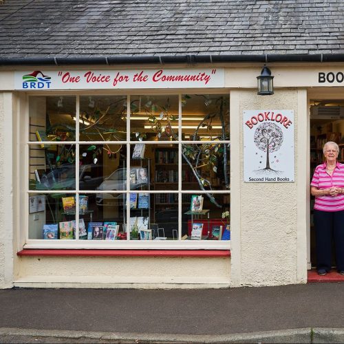 A woman in a pink shirt stands in a doorway next to a large window displaying books decorated with tree branches in a shop.