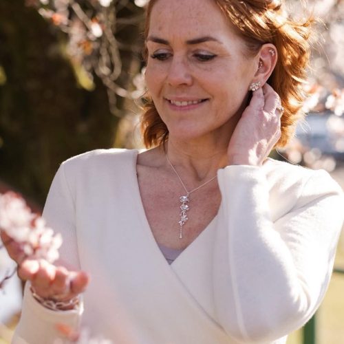 A smiling woman wearing a white shirt and a necklace with a silver chain and floral pendant paired with matching floral earrings.