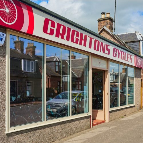 Crightons Cycles has six large windows displaying bikes underneath a bright pink and white sign.