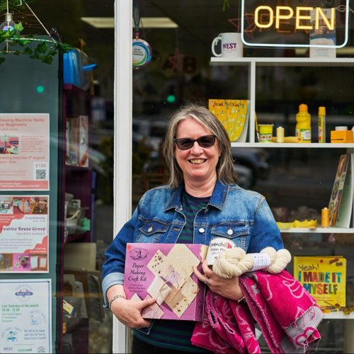 A smiling woman wearing sunglasses and a blue jacket holds pink and beige craft supplies and a pink blanket outside a shop