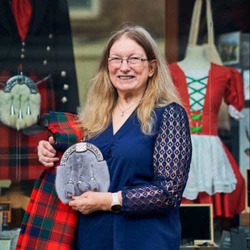 A smiling woman wearing a blue shirt holds a red kilt and grey sporran outside a shop window displaying other Scottish items.