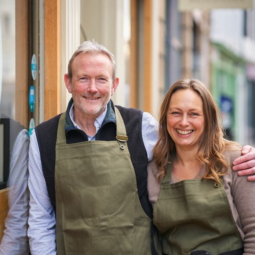 A smiling man rests his hand on a smiling woman's shoulder, both wearing a khaki apron