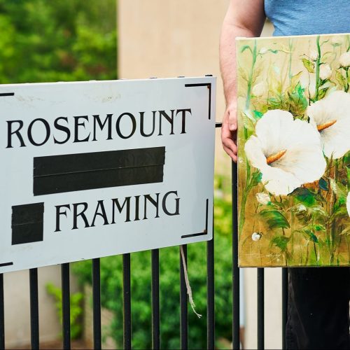 A white sign with black writing mounted on a fence next to a painting of flowers held by a person.