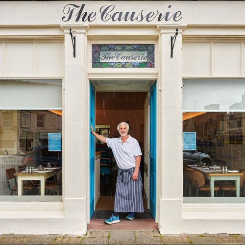 An older man wearing a grey shirt and stripped apron leans on a blue door outside The Causerie