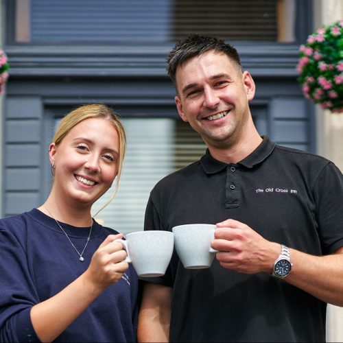 A smiling woman in a blue shirt and a smiling man in a black shirt stand next to each other holding mugs in front of a window.