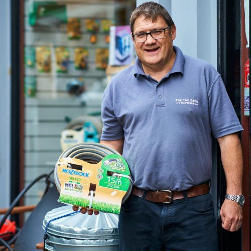 A smiling man in a grey shirt holds a garden hose coiled up in packaging while standing in front of a hardware store.