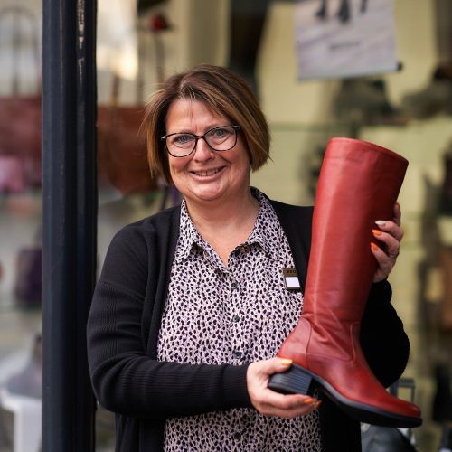 A smiling woman wearing glasses and a patterned shirt holds a red women's boot in a shop doorway