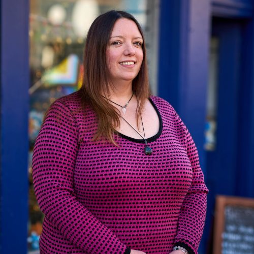A smiling woman with brown hair wearing a pink and black patterned shirt stands outside a shop painted blue