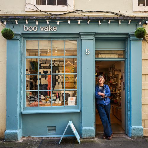 A woman wearing a stripped shirt leans against a doorway painted blue next to a large window displaying textiles and small gifts