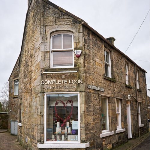 A stone building on a street corner features a salon with a large window displaying hair products and decorative hearts