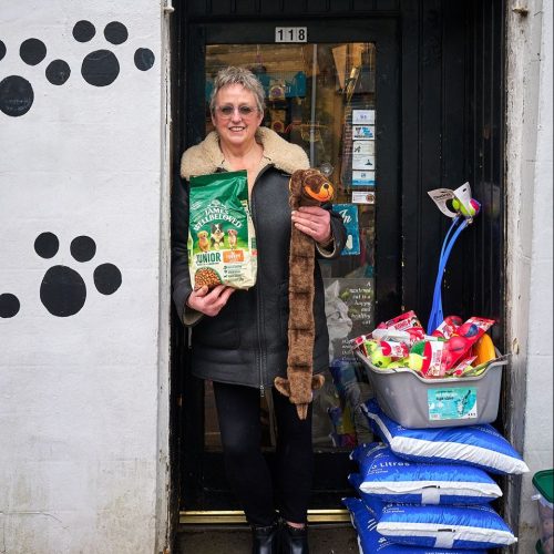 A smiling woman wearing a black jacket stands in a doorway decorated with paw prints holding a bag of dog food and a long dog toy