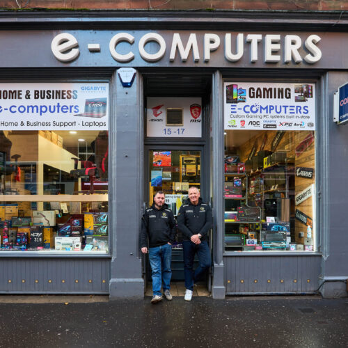 Two smiling men wearing matching black jackets stand outside a grey shop with two large windows displaying computer equipment