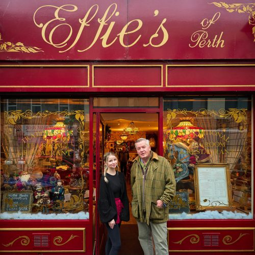 A young woman wearing black stands next to an older man wearing green outside a cafe painted red with two decorated windows