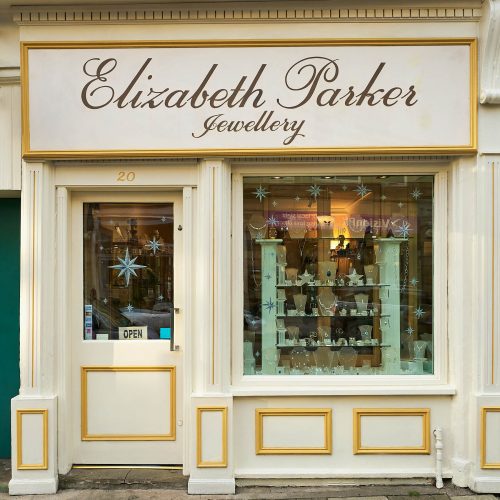 A white shop has a sign reading Elizabeth Parker Jewellery written in cursive above a window displaying an array of jewellery