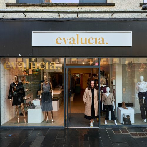 A smiling woman wearing a beige coat stands outside a shop with two large windows displaying dressed golden mannequins