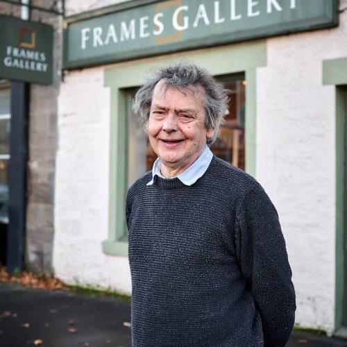 An older man with grey hair wearing a grey jumper stands outside a white shop with a large green sign