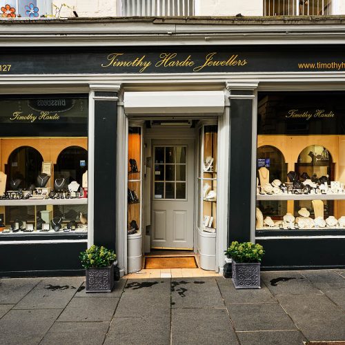 A black and white shop front with signs written in gold cursive font and two large windows displaying a collection of fine jewellery