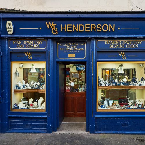 A blue shop in Perth with signs written in gold font above two large windows displaying an array of jewellery either side of a door