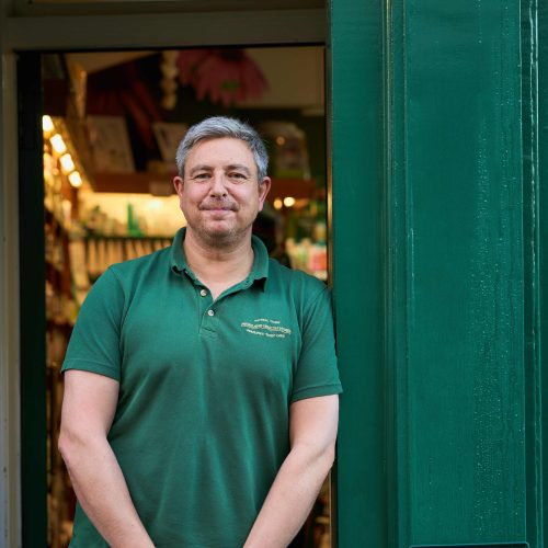 A man with grey hair wearing a green shirt leans against a doorway painted the same shade of green outside a shop