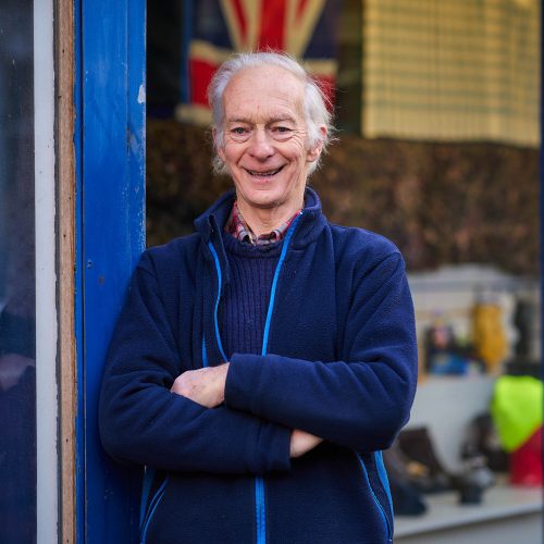 A smiling older man wearing a blue jumper has his arms crossed and stands in a doorway outside a shop