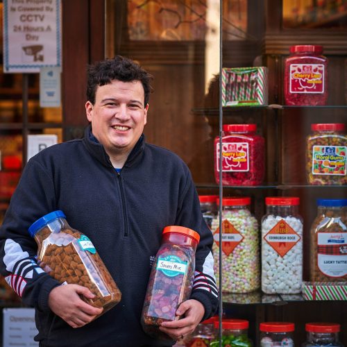 A smiling man wearing a black shirt holds two jars of candy outside a window displaying an assortment of similar jars