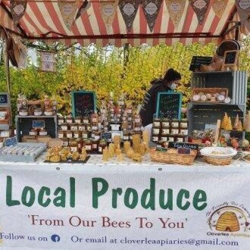 A stall with a stripped canopy and a large sign reading Local Produce displays a collection of jars of honey and honey comb