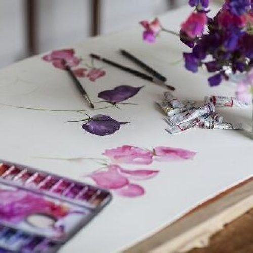 A pink and purple watercolour painting of flowers sits next to a paint pallet and a jar of pink and purple flowers