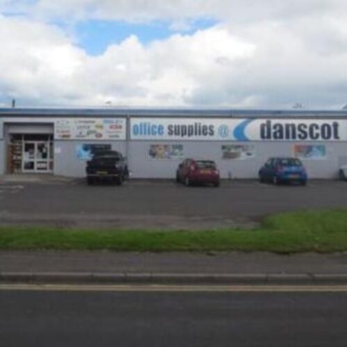 A short white building with a large white sign reading Danscot office supplies sits in a car park populated by four cars