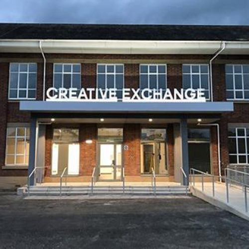 A large stone building with several windows has a large entry way with a white sign reading Creative Exchange above the doors