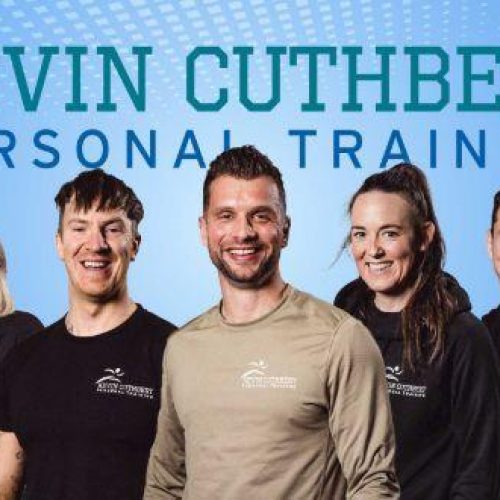 Four smiling people wearing black shirts stand either side of a smiling man wearing a beige shirt in front of a blue background