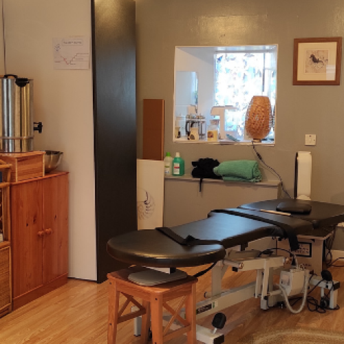A long black table sits in a cluttered room next to a  wooden cabinet, a black cupboard, a window, and a mirror