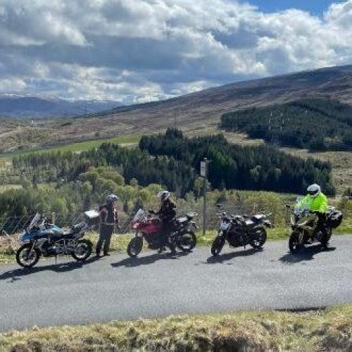 Five motorcycles are parked in a line in on a mountainous country road, with three riders gathered around them