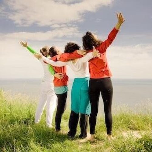 A group of four people wearing brightly coloured clothes stand arm in arm on a grassy hill looking out over a large body of water