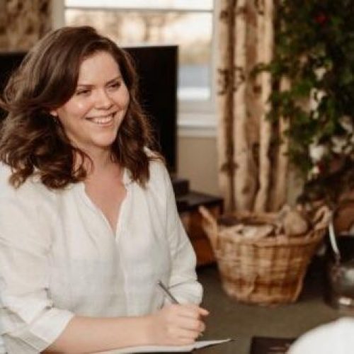 A smiling woman wearing a white shirt holds a pen and paper and sits across from a person with brown hair