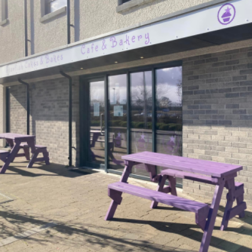 A stone building with glass sliding doors has a purple and white sign and purple benches sitting outside