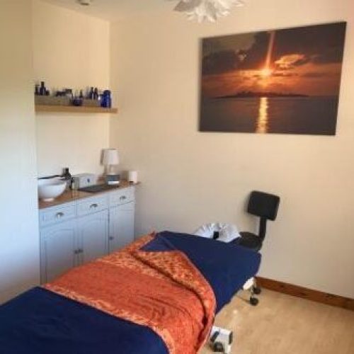A blue massage bed draped with an orange blanket sits in a white room decorated with a cabinet, a shelf, and a painting of a sun set