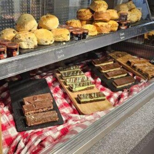A small shelf full of various flavours of scones and jam sits above a gingham counter with a collection of different desserts